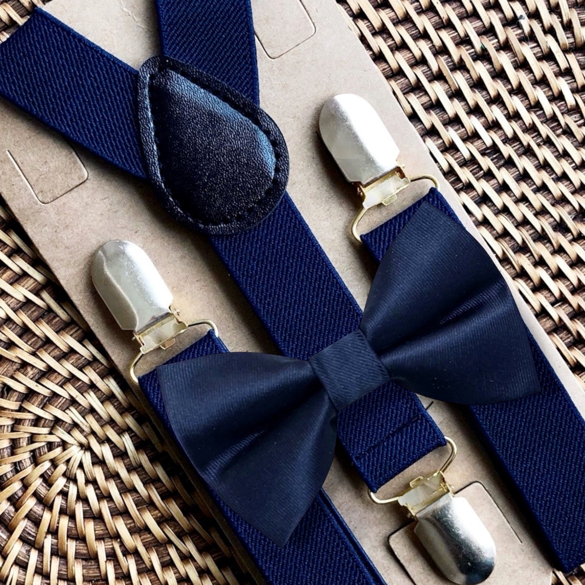 Navy bow tie and navy blue suspenders with silver clips and a matching bow tie on a woven surface.