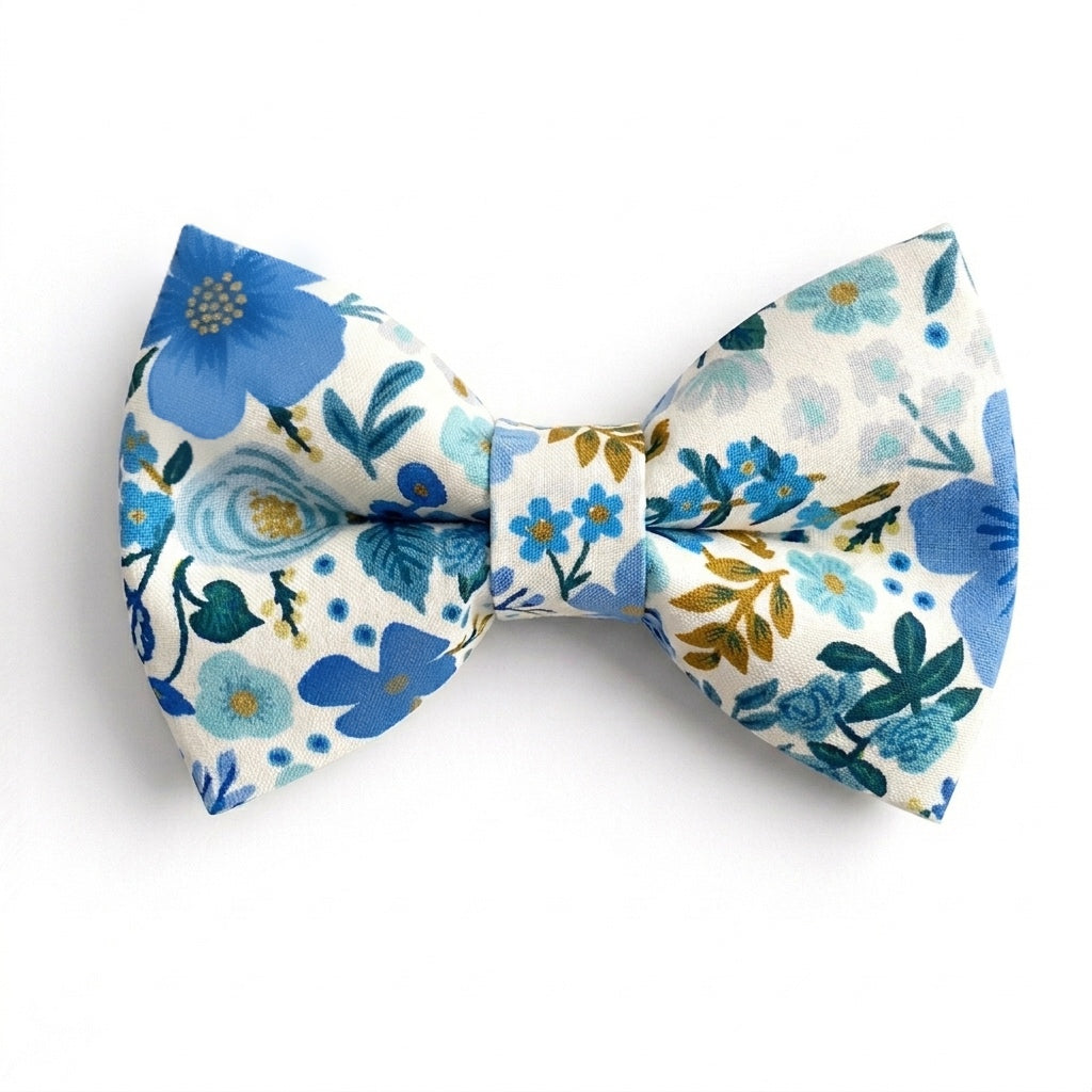 A pre-tied bow tie featuring a cream background with a vibrant pattern of light and dark blue flowers, green foliage, and gold accents.