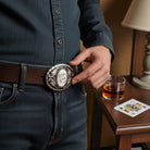 Person adjusting a belt with a decorative cowboy deck of cards belt buckle, sitting at a table with a glass of whiskey and playing cards.