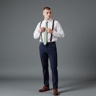 Groomsman wearing a white shirt, adjusting a sage tie, and dark brown suspenders for a wedding party.