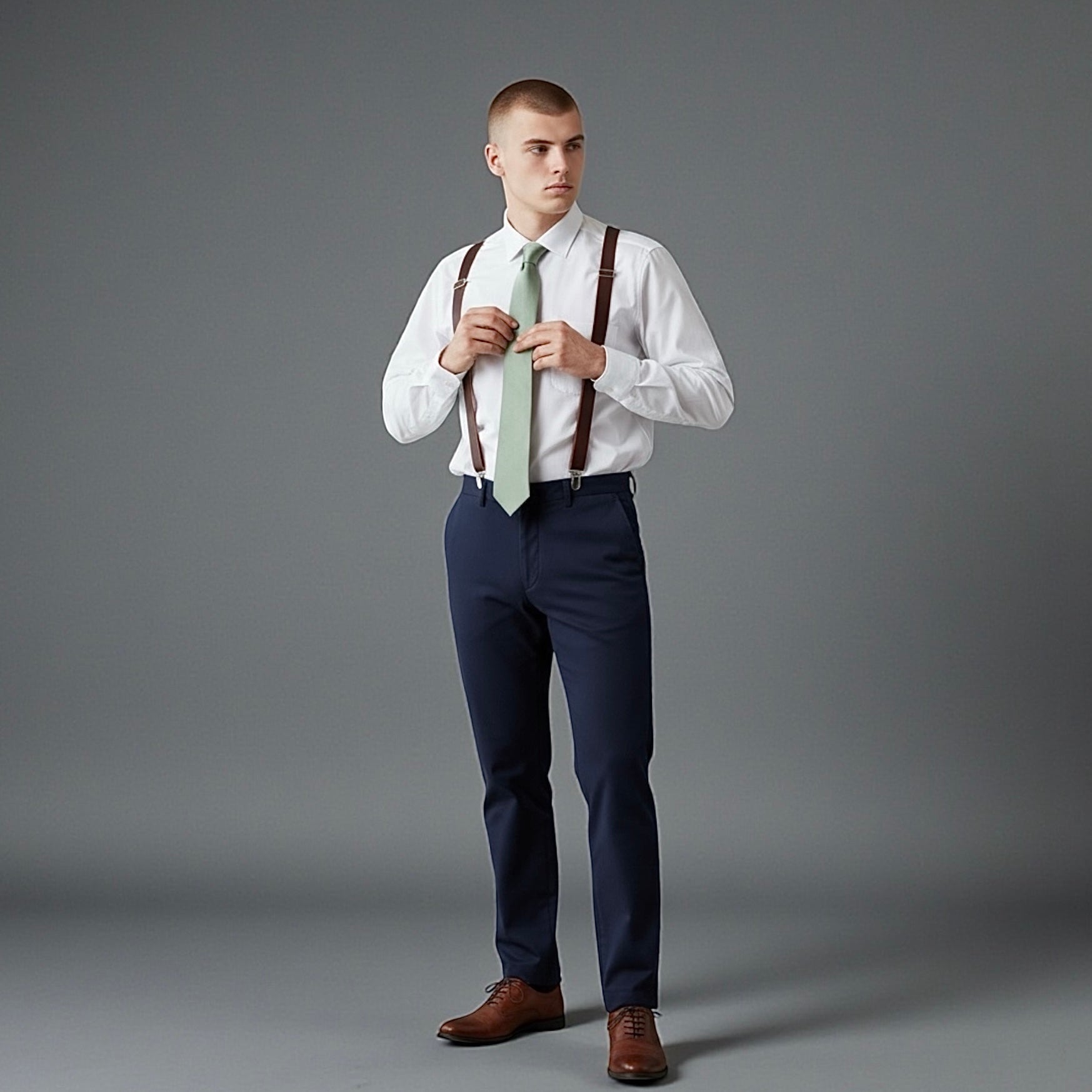 Groomsman wearing a white shirt, adjusting a sage tie, and dark brown suspenders for a wedding party.