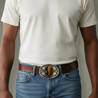 Man wearing a white t-shirt, blue jeans, and a brown belt with a decorative tiger’s eye belt buckle.