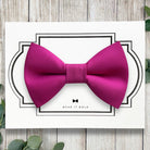 Pink dog bow tie on a white card with decorative border and 'Wear It Bold' text, placed on a wooden surface.