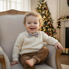 Child wearing a Christmas bow tie sitting on a chair with a decorated Christmas tree in the background