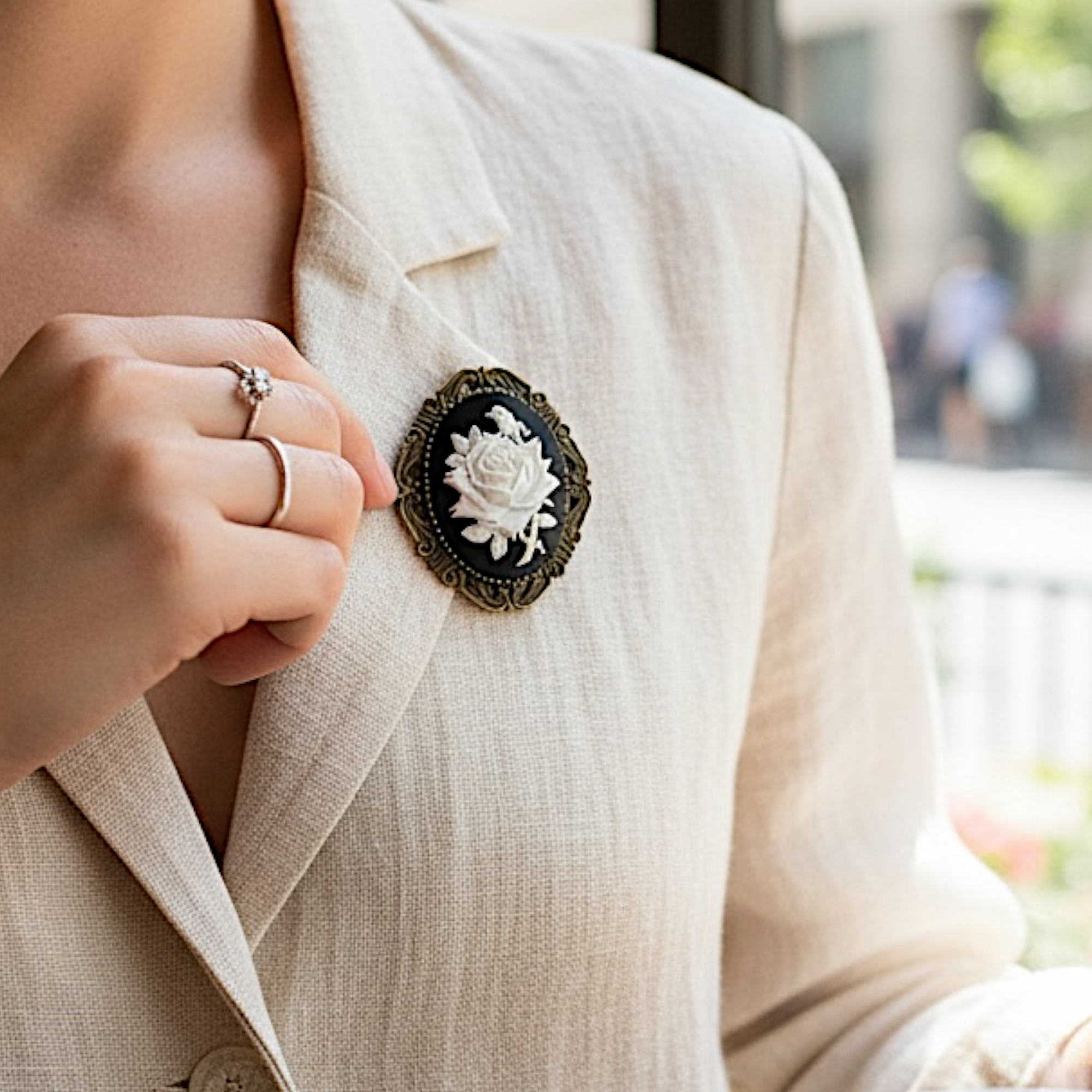 Person wearing a beige blazer with a decorative flower brooch and rings on fingers.