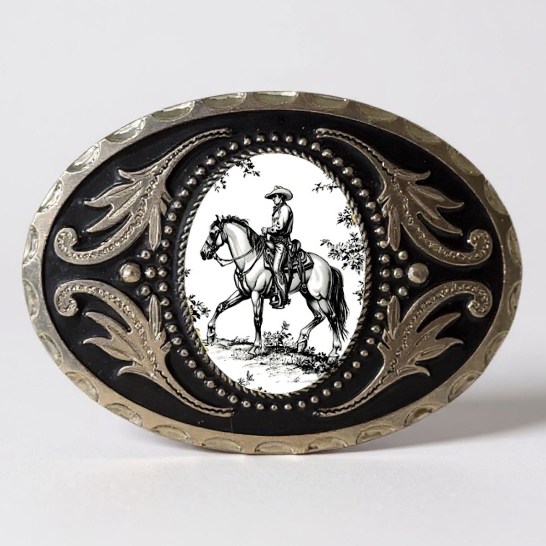 Decorative Cowboy belt buckle with a black background and white illustration of a person on horseback, on a light gray background.