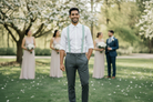 A groomsman wearing sage green suspenders with leather accents and a white dress shirt standing at an outdoor forest wedding.