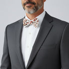Man wearing a gray suit with a white shirt and patterned burnt orange floral bow tie on a light gray background