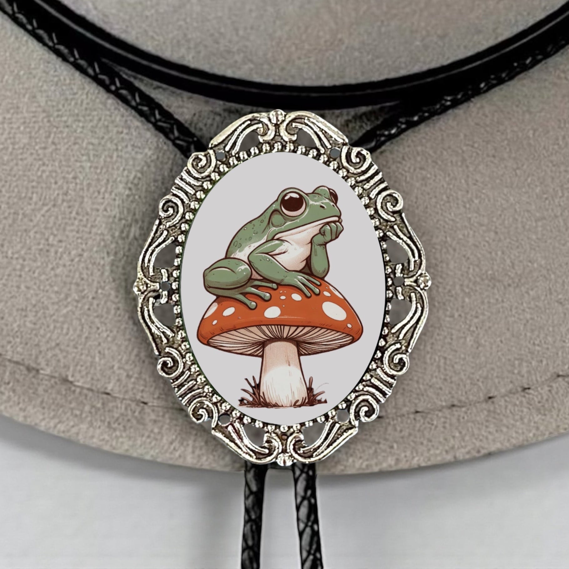 Cottagecore bolo tie with a frog on a mushroom design on a leather strap against a beige background