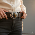 Person wearing a brown tiger’s eye belt buckle with an ornate buckle, holding it with both hands.