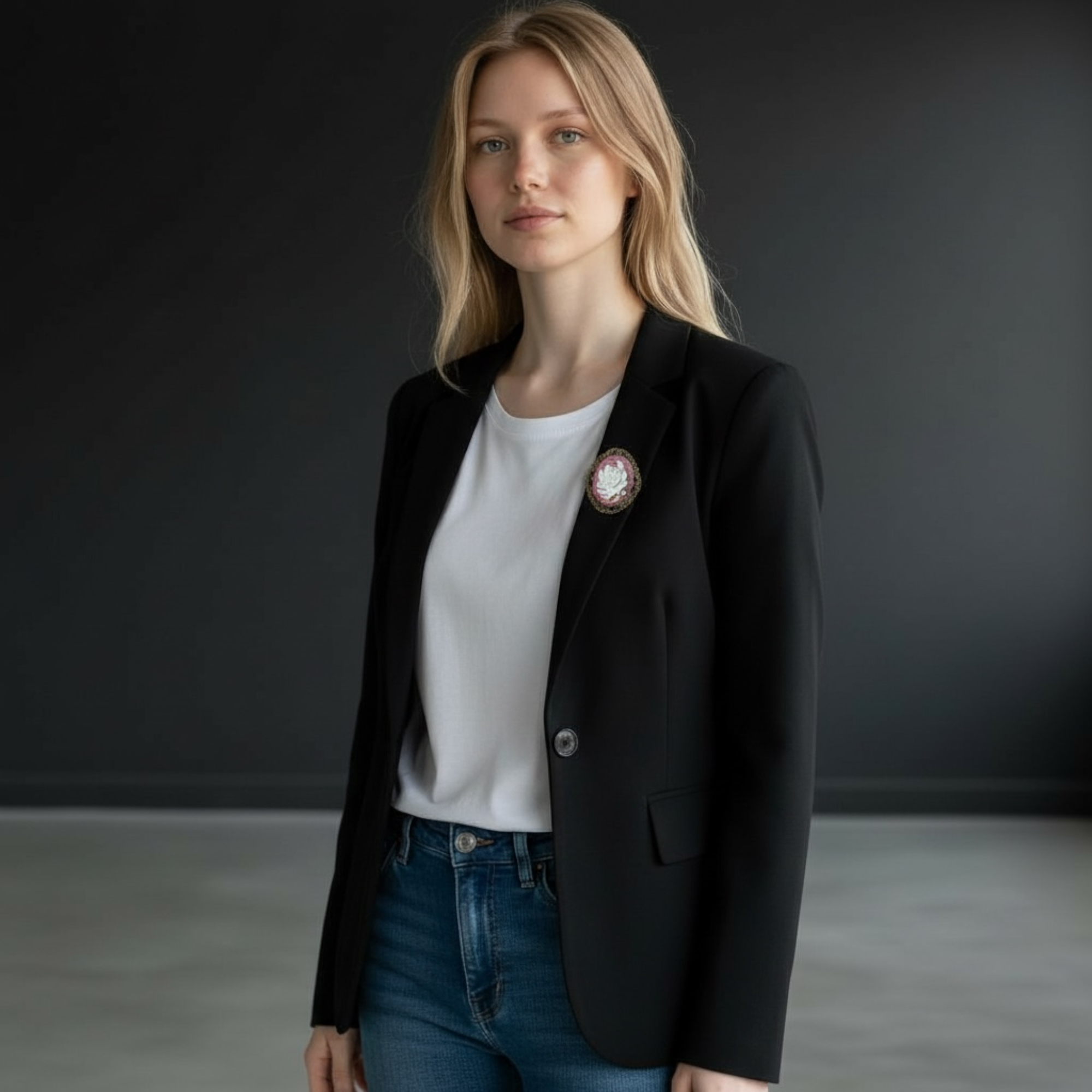 Woman wearing a black blazer over a white shirt with a pink floral brooch, standing against a dark background.