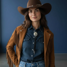 Woman wearing a brown cowboy hat, raven bolo tie, fringed jacket, and blue denim shirt against a dark blue background.