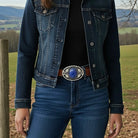 Person wearing a denim jacket and jeans with a decorative belt buckle with blue stone in a natural setting.