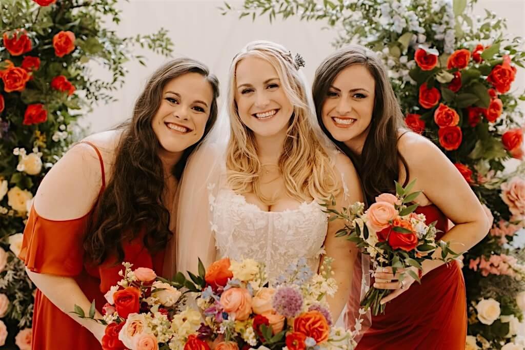 Two bridesmaids and a bride at a garden wedding two in red dresses and one in a white wedding dress, standing in front of floral decorations. These are the sisters who hand make the items together at The Bold Bow Tie.