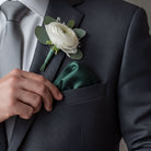 A close-up of a man's hand holding a green napkin in his suit jacket pocket, with a white rose boutonniere pinned to the lapel.