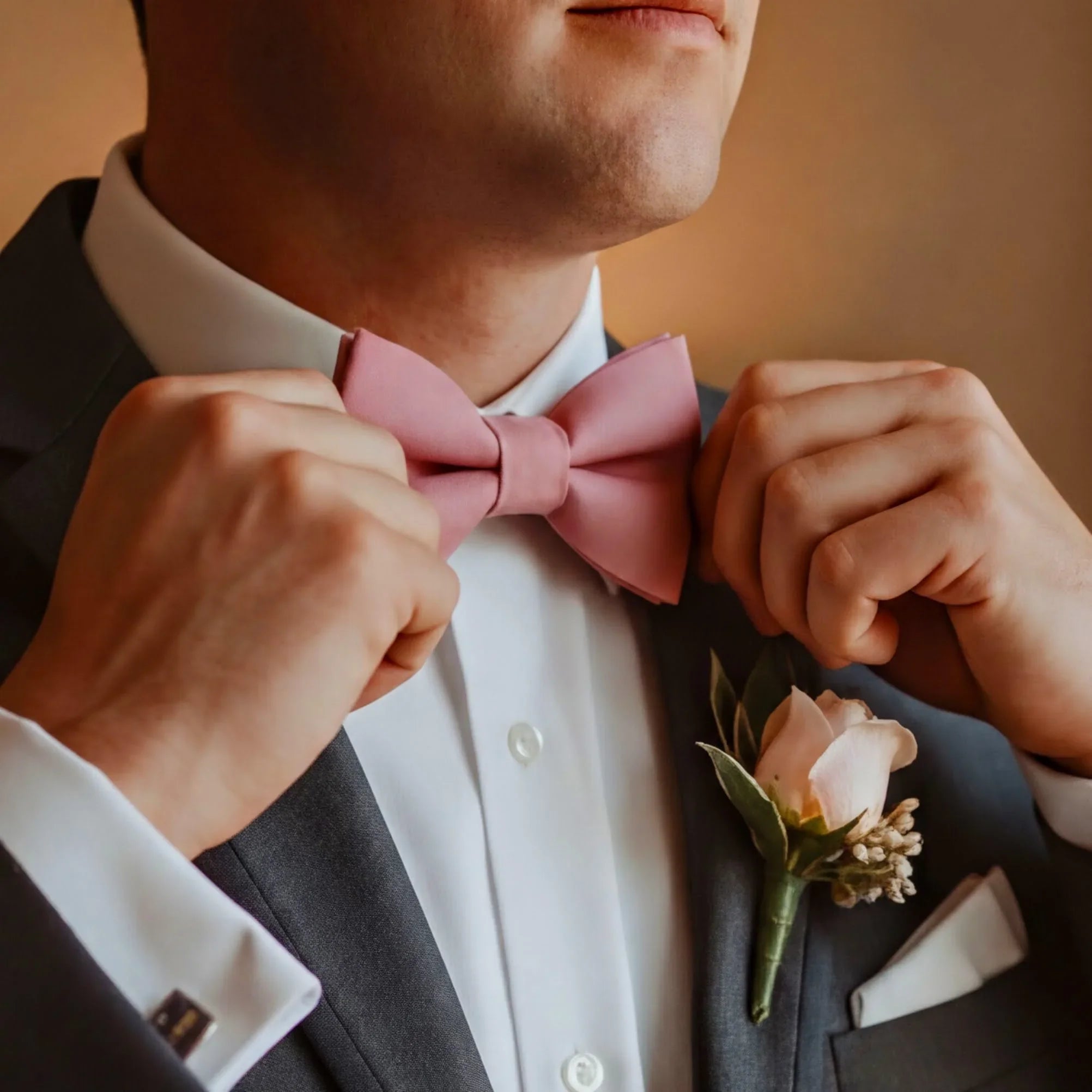 A man in a suit and pink bowtie is adjusting his tie, with a boutonniere pinned to his lapel.