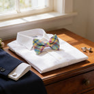 A white dress shirt with a colorful plaid bow tie is displayed on a wooden dresser, accompanied by a pair of cufflinks and a jacket.