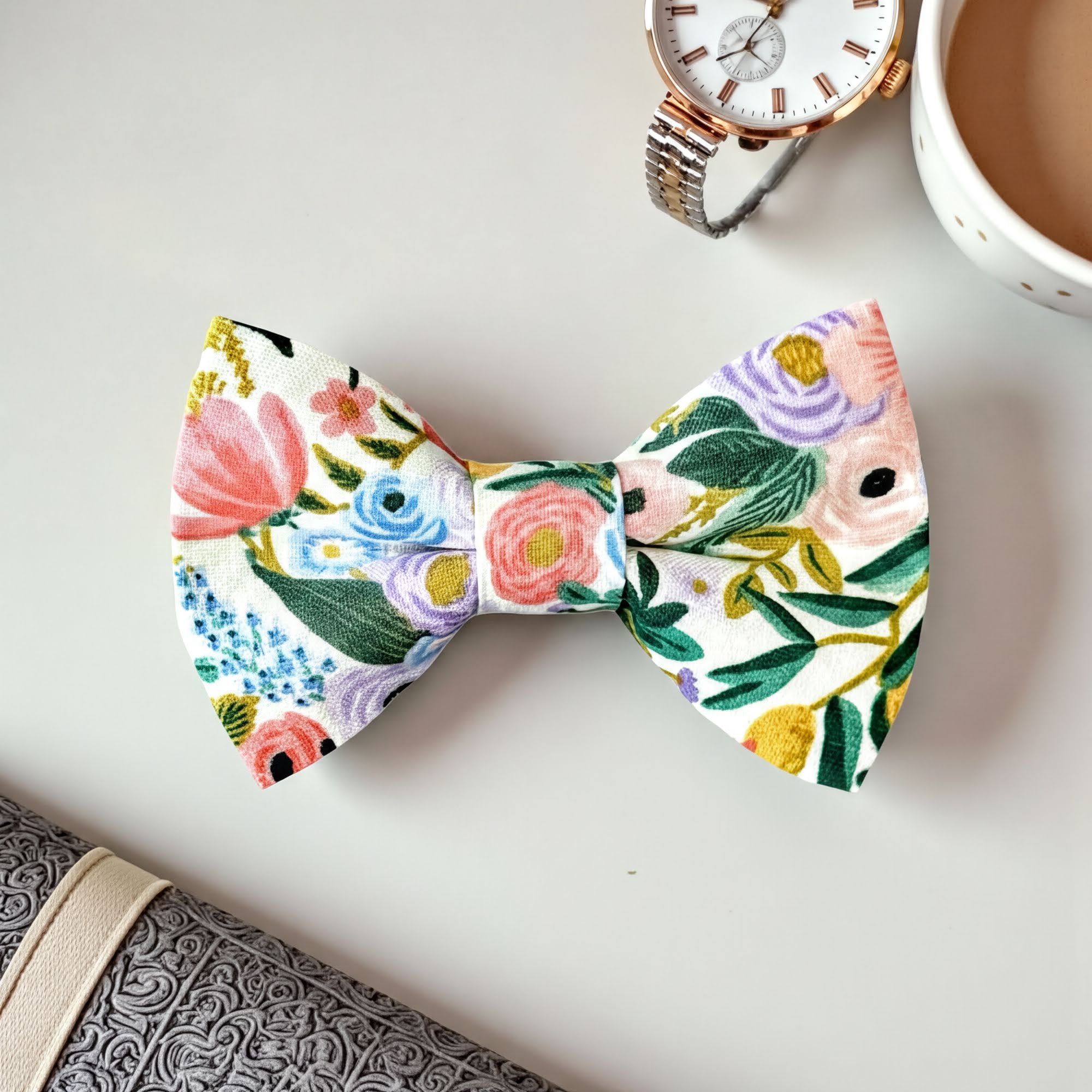 Floral bow tie on a white surface with a watch and cup of coffee in the background