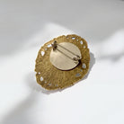A decorative pin with a gold leaf-like design and a round center, placed on a white surface.