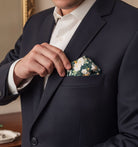 A groomsman in a dark suit jacket is holding a pocket square with a floral pattern.