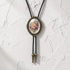 A decorative floral pendant necklace with a black cord and gold-toned metal frame is displayed against a white textured wall.