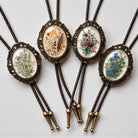 Handmade wedding floral bolo ties with vintage bronze oval frames, braided leather cords, and botanical flower designs for groom, groomsmen, rustic weddings, western wedding attire, and floral formal accessories.