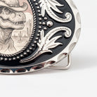 A decorative metal buckle featuring a detailed illustration of a dinosaur wearing a cowboy hat, with a desert landscape in the background.