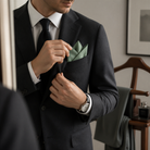 A man in a black suit jacket is adjusting his pocket square, with a watch visible on his wrist.