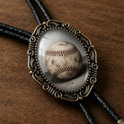A baseball bolo tie for men and women displayed in a decorative oval-shaped frame on a wooden surface.