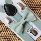 Sage green bowtie and suspenders for a wedding on a woven surface.