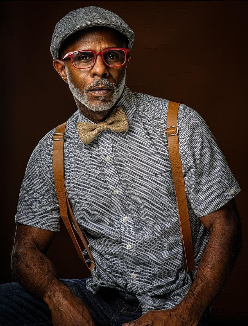 A handsome man in red glasses and a page boy hat wears a country chic burlap bow tie and rustic tan suspenders for men and boys to wear to a cowboy wedding.